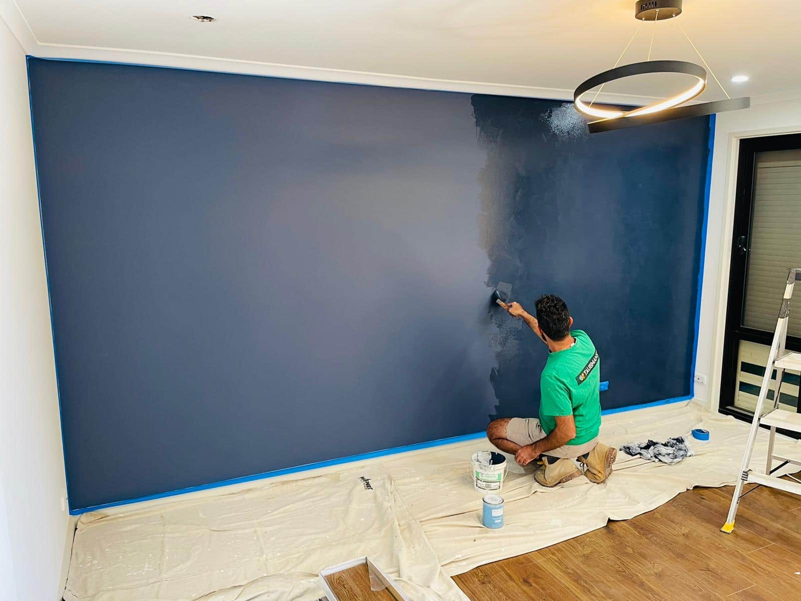 Painter applying Porter’s French Wash to a blue feature wall in St Clair, with edges masked and timber floor protected