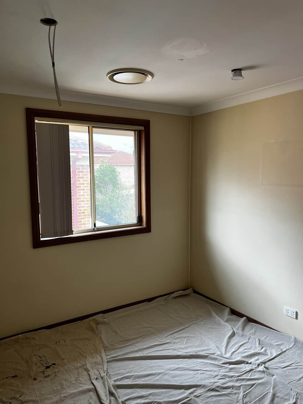 Beige bedroom walls beneath a white ceiling, with drop sheets covering the floor before painting.