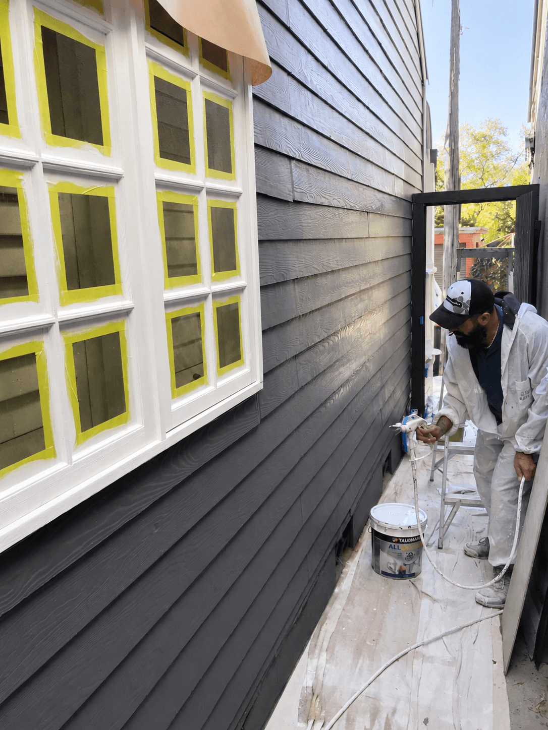 Painter spray painting exterior weatherboards beside masked windows in Stanmore